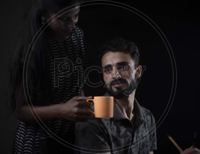 Young brunette Indian/African dark skinned lady offering coffee/tea to her European/Arabian/Kashmiri man/husband at his work in black copy space studio background. Lifestyle of a couple.