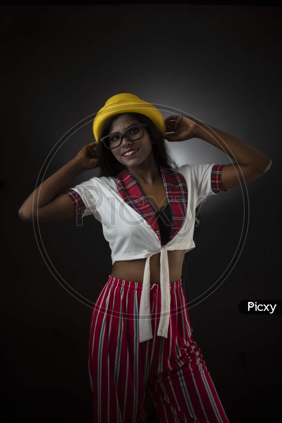 Portrait of an Asian/Indian/African brunette dark skinned young girl in sexy school uniform and spectacles with yellow hat in a black/grey studio background. Fashion and cosplay photography.