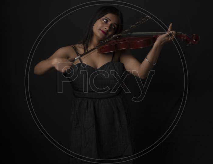 Fashion portrait of an young and attractive Indian Bengali brunette girl with black western dress is playing violin in front of a black studio background. Indian fashion portrait and lifestyle.