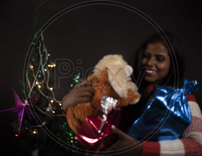 Portrait of young and attractive brunette dark skinned Indian Bengali woman in western woolen sweater with soft toy in decorative Christmas tree background.Indian lifestyle and Christmas celebration