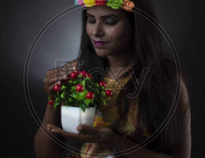 Fashion portrait of young dark skinned Indian brunette girl in floral dress with head band holding cherry tree in front of black copy space studio background. Indian fashion portrait and lifestyle.