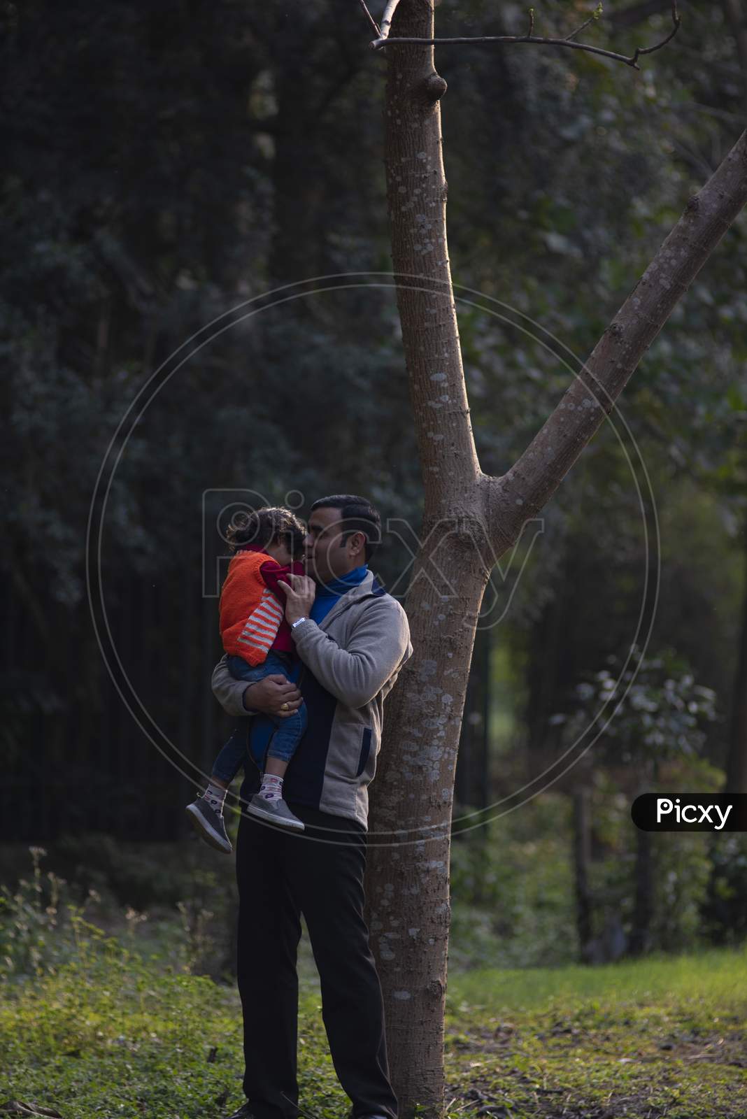 An Indian brunette father and his baby boy in winter garments enjoying themselves in winter afternoon on a  green grass field in forest background. Indian lifestyle and parenthood.