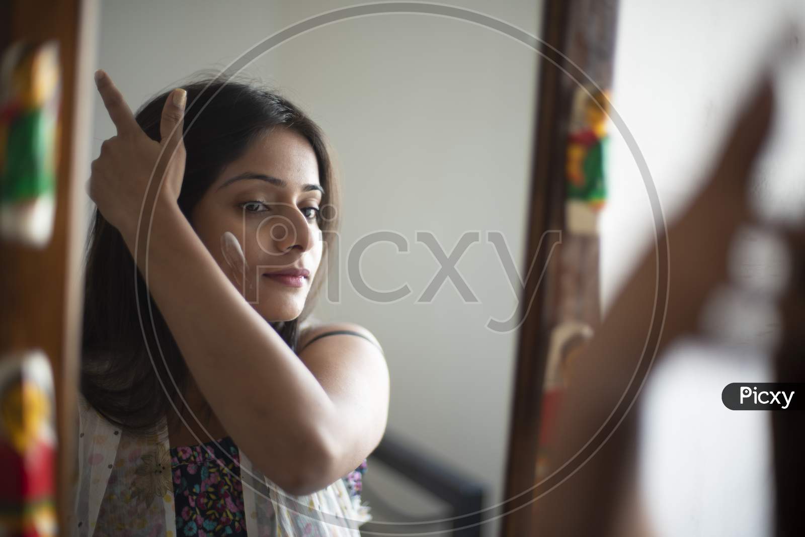 Reflection of an young and attractive Indian Bengali brunette woman is looking her face in a mirror while applying a white beauty cream on her face in her room. Indian lifestyle.