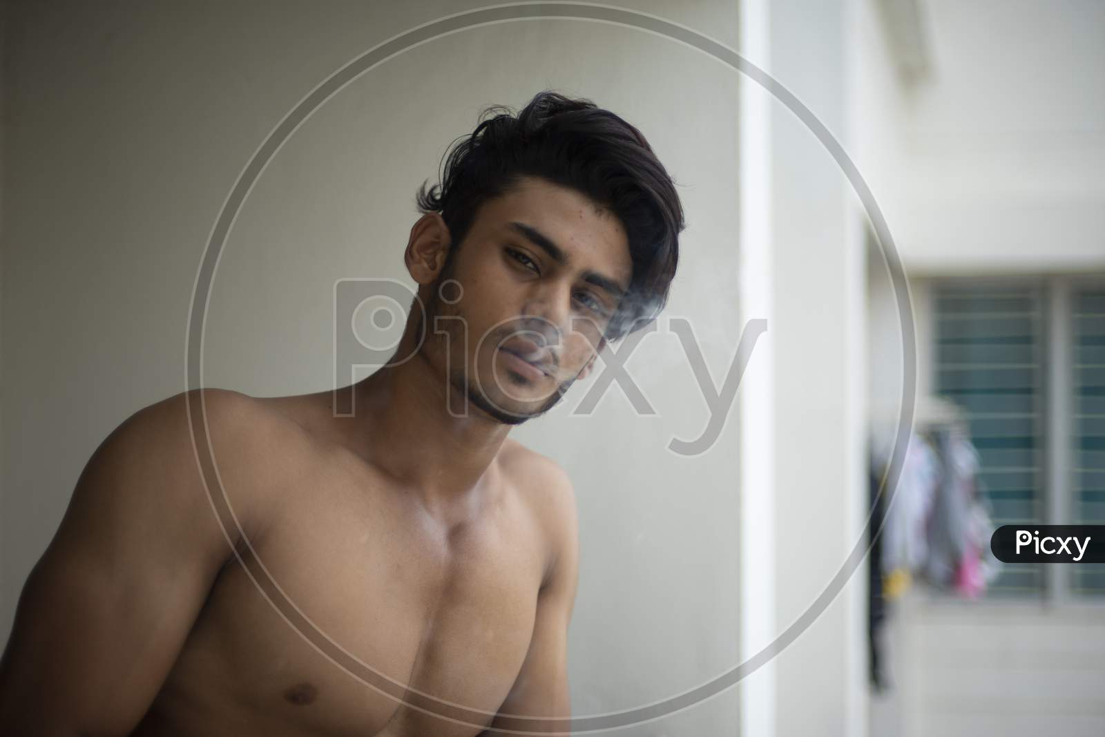 Image of Portrait of an young and handsome brunette Bengali muscular man in  bare body smoking on a balcony in white urban background. Indian  lifestyle.-CZ620182-Picxy