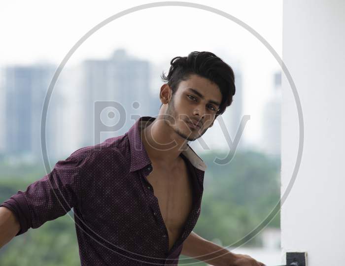 Portrait of an young and handsome brunette Bengali muscular man in casual shirt and jeans standing on a balcony in white urban background. Indian lifestyle.