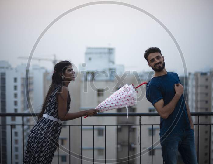 A dark skinned Indian/African girl in  western dress and a Kashmiri/European/Arabian man in casual wear having fun with umbrella on a rooftop in urban background. Indian lifestyle.