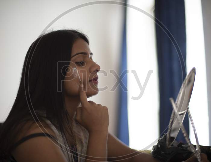 Side portrait of an young and attractive Indian Bengali brunette woman is looking her face in mirror while applying a white beauty cream on her face in her room in white background. Indian lifestyle.
