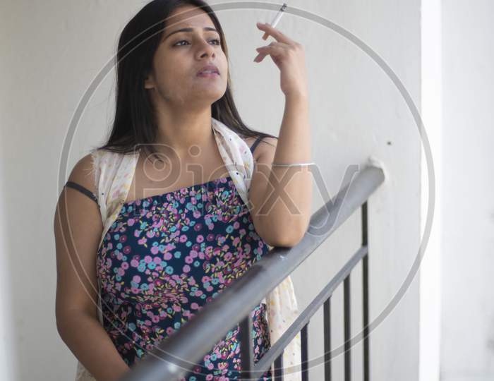 An young and attractive Indian Bengali brunette woman in white sleeping wear is smoking cigarette on the balcony in the morning in white background. Indian lifestyle.