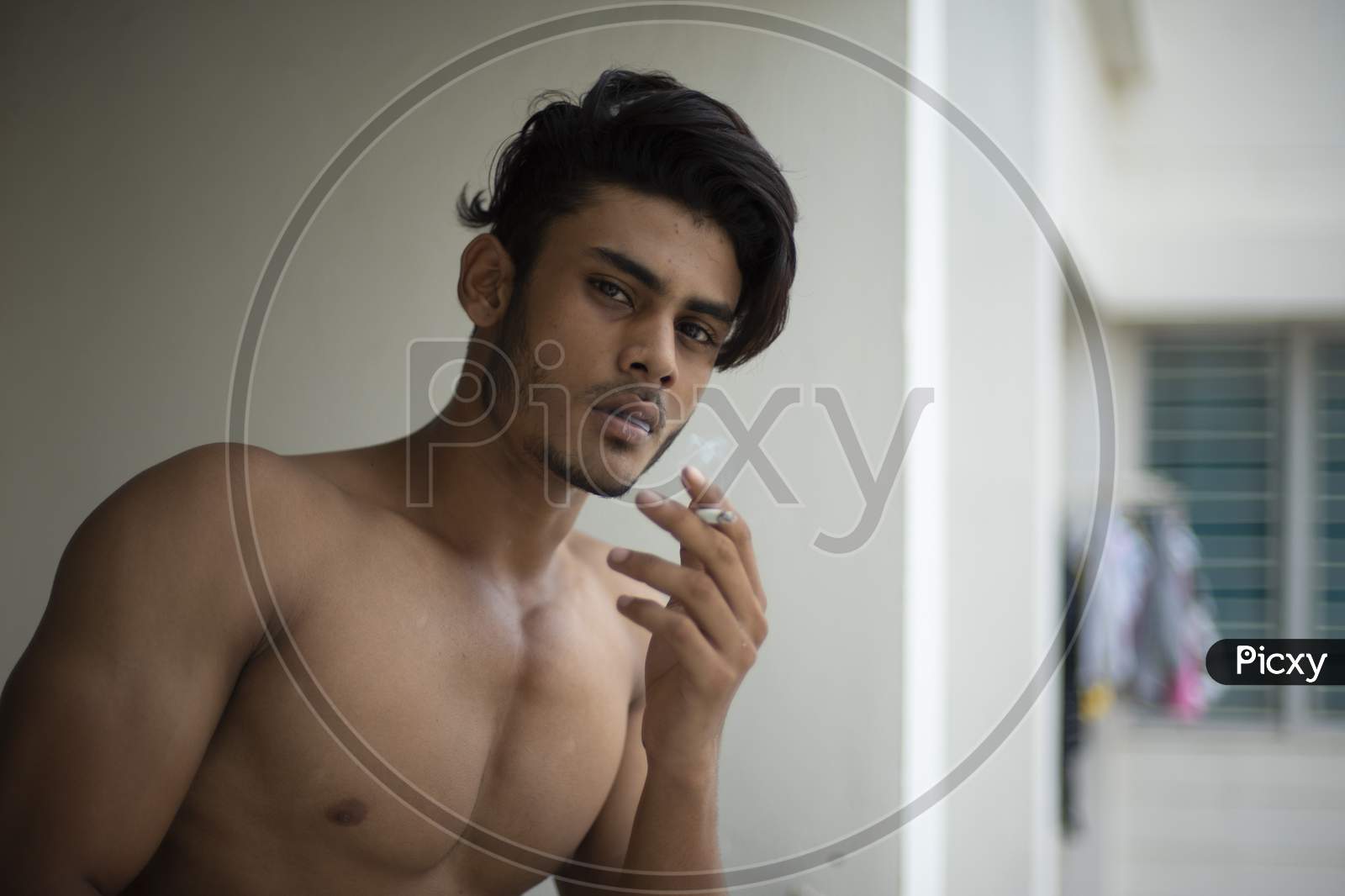 Portrait of an young and handsome brunette Bengali muscular man in bare body smoking on a balcony in white urban background. Indian lifestyle.