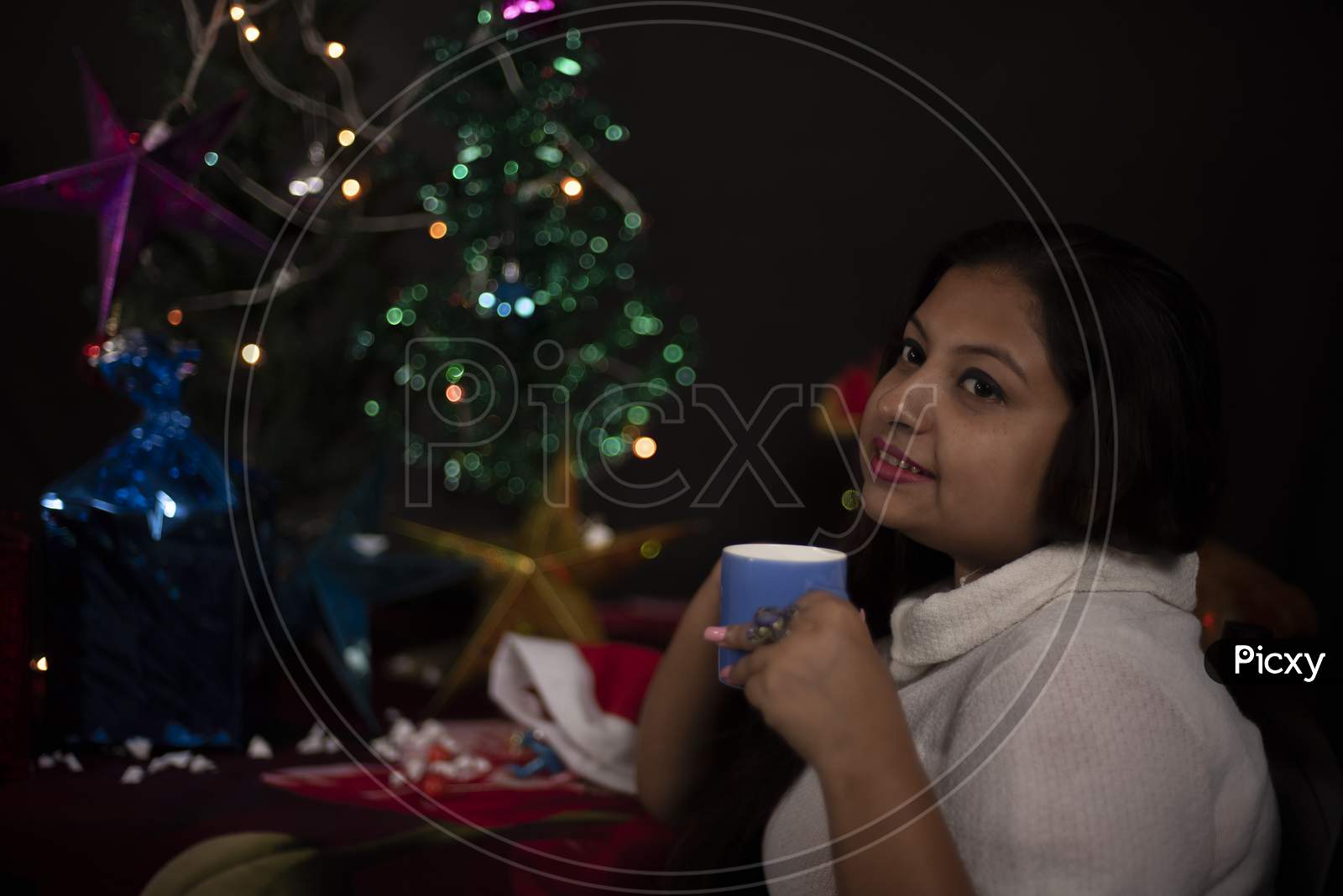 An young and attractive brunette plus size Indian Bengali woman in western woolen sweater enjoying with coffee/tea in decorative Christmas tree background.Indian lifestyle and Christmas celebration