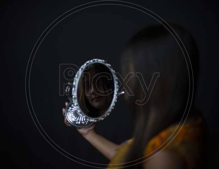 Reflection of an young and attractive Indian Bengali brunette woman is looking her face in a mirror while doing makeup in black studio copy space background.Indian lifestyle.