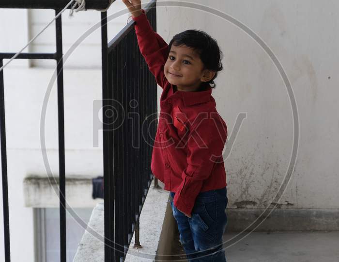 Portrait of Indian cute little cheerful brunette Tamil baby boy wearing vibrant red shirt while standing on a balcony in a white urban background. Indian lifestyle.
