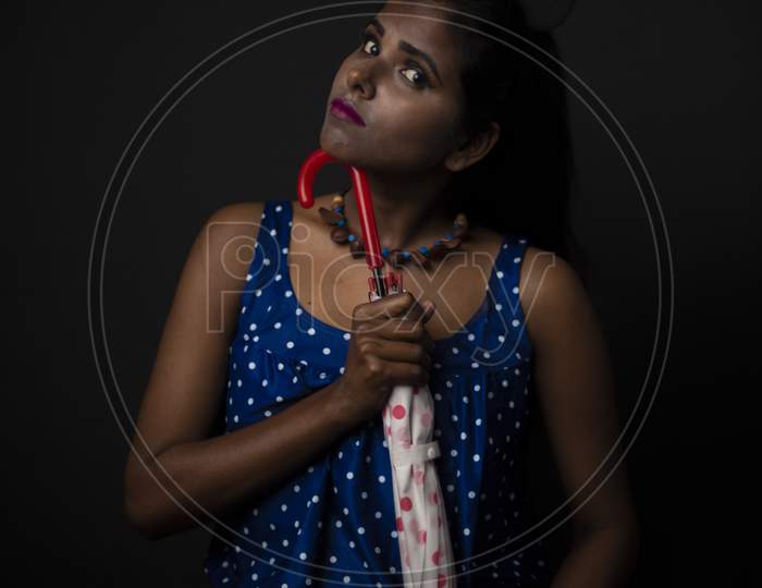 Fashion portrait of young dark skinned Indian/African brunette girl in blue and red western dress holding an umbrella in front of black copy space studio background. Indian fashion photography.