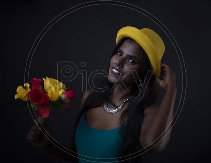 Fashion portrait of young dark skinned Indian/African brunette girl in green western dress and yellow hat holding flowers in front of black copy space studio background. Indian fashion photography.