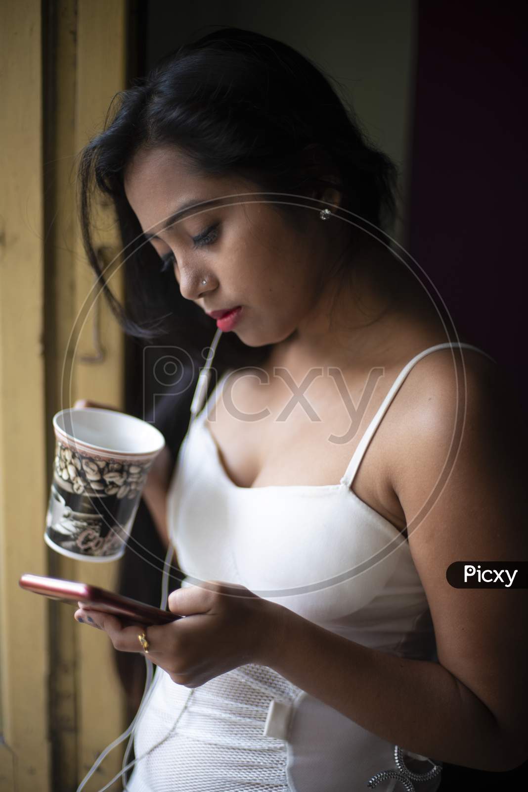An young and attractive Indian  brunette woman in white sleeping wear standing with a coffee/tea cup and cellphone in front of a window  inside in her room. Indian lifestyle.