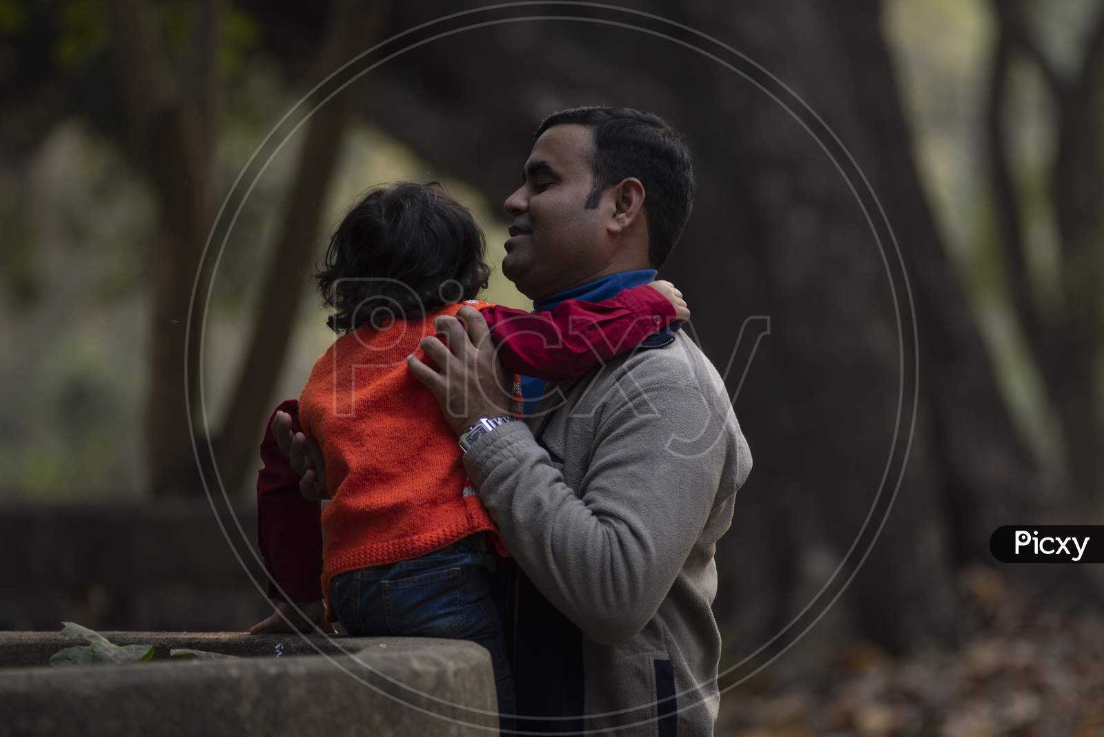 An Indian brunette father and his baby boy in winter garments enjoying themselves in winter afternoon on a  green grass field in forest background. Indian lifestyle and parenthood.