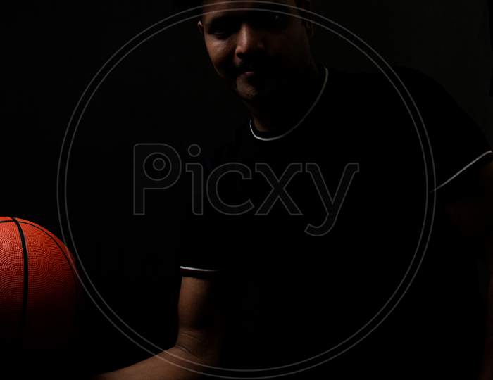Portrait of a handsome and intelligent Indian brunette man wearing a solid black t shirt standing before a copy space black background holding a basketball. Indian lifestyle and fashion portrait