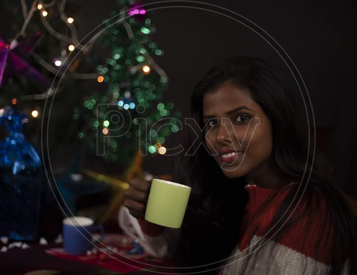 An young and attractive brunette dark skinned Indian Bengali woman in western woolen sweater enjoying with coffee/tea in decorative Christmas tree background.Indian lifestyle and Christmas celebration