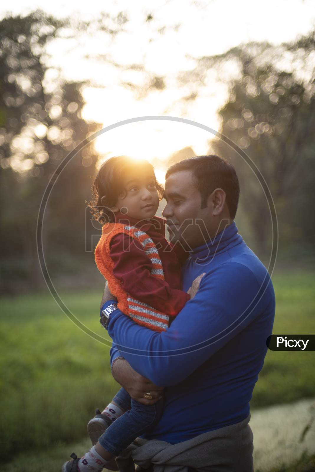 An Indian Telugu brunette father and his baby boy in winter garments enjoying themselves in winter afternoon on a  green grass field in forest background. Indian lifestyle and back light photography.