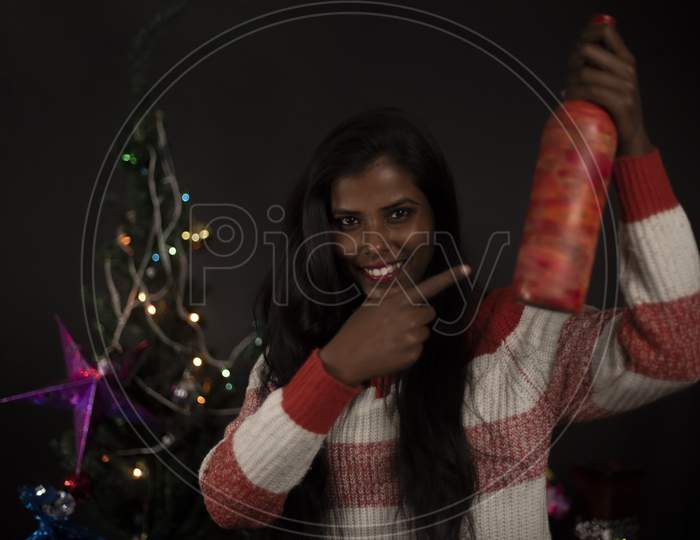 Portrait of an young and attractive brunette dark skinned Indian Bengali woman in western woolen sweater showing gift in decorative Christmas tree background.Indian lifestyle and Christmas celebration