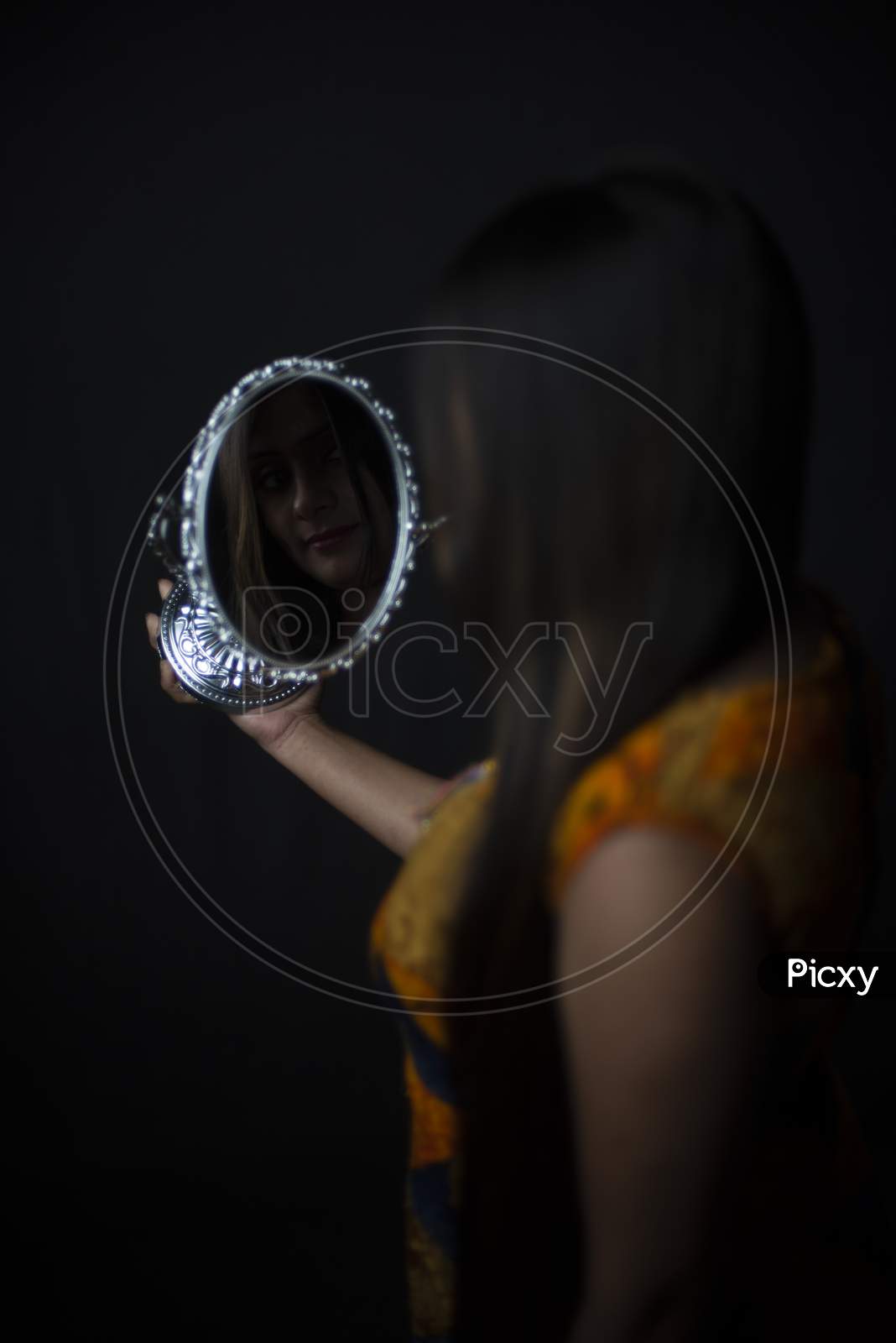 Reflection of an young and attractive Indian Bengali brunette woman is looking her face in a mirror while doing makeup in black studio copy space background.Indian lifestyle.