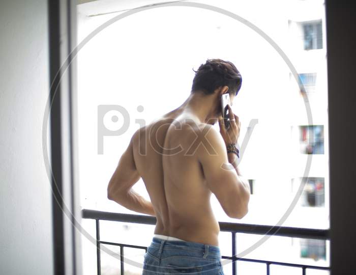 Portrait of an young and handsome brunette Bengali muscular man in bare body standing on a balcony with phone in white urban background. Indian lifestyle.