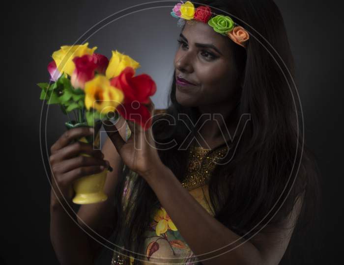 Fashion portrait of young dark skinned Indian Bengali brunette girl in floral dress with floral head band taking flowers in front of black copy space studio background. Indian fashion photography.
