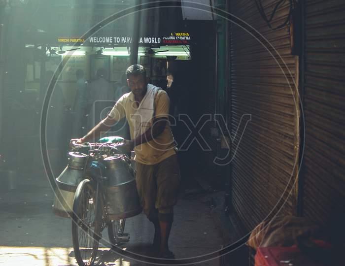 New Delhi, Delhi India- May 31 2020: A Milk Vendor Cycling Through Narrow Streets In Delhi In The Morning, Beautiful Tyndall Effect By The Sun Rays In The Morning.
