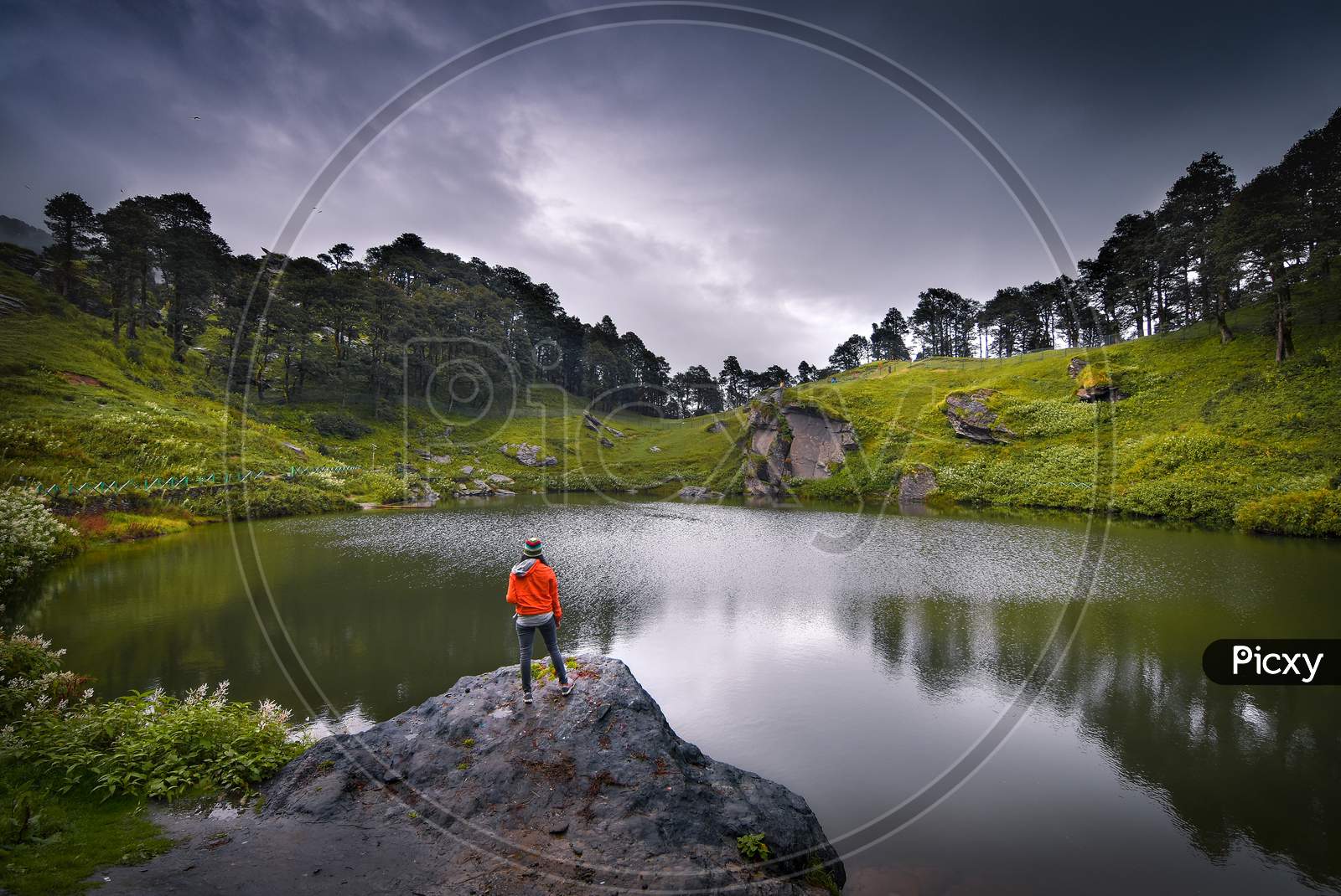 A Landscape Of A Small Lake In Between The Mountain Ranges With A Man Standing Beside It.