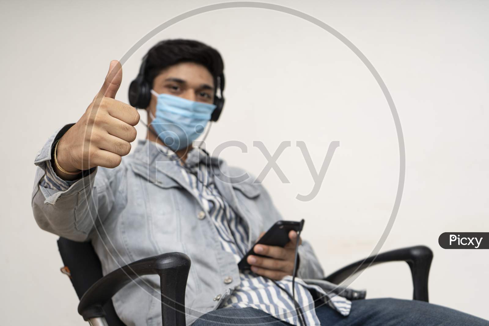 Young Handsome Asian Indian Boy Sitting On A Chair, Wearing A Protective Mask, Showing Thumbs Up In The Camera.