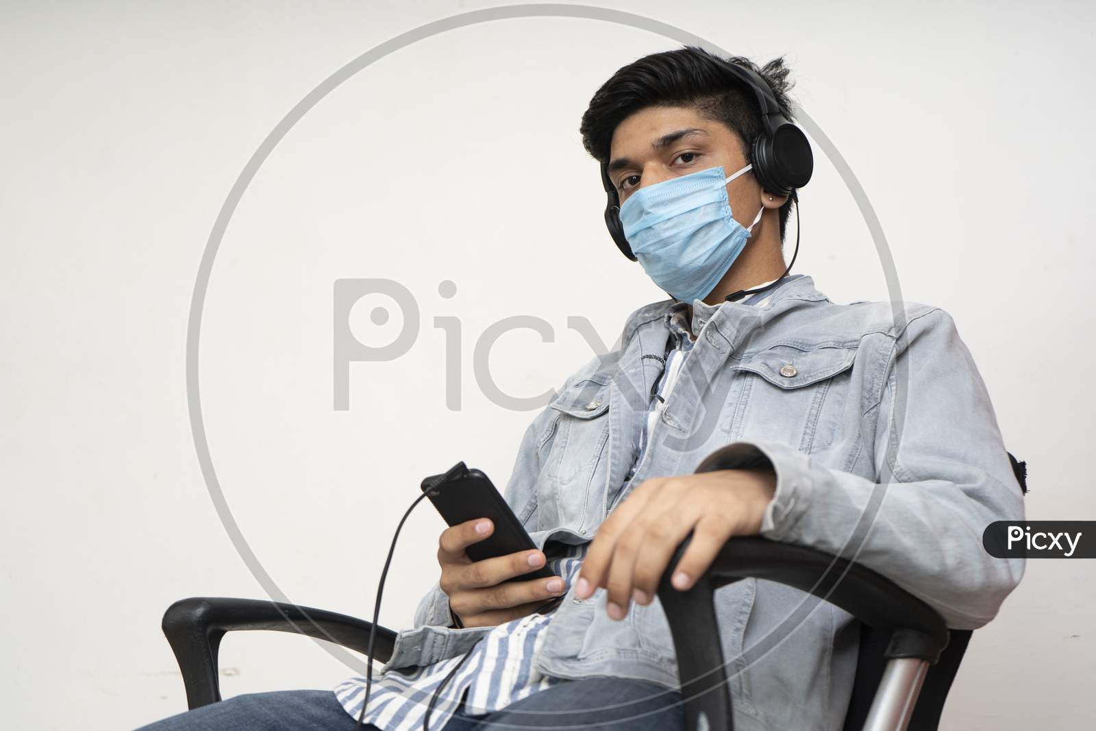 Young Handsome Asian College Going Boy Sitting On A Chair, Wearing A Protective Mask, Listening To Songs On His Headphones.