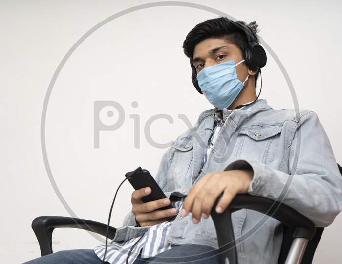 Young Handsome Asian College Going Boy Sitting On A Chair, Wearing A Protective Mask, Listening To Songs On His Headphones.