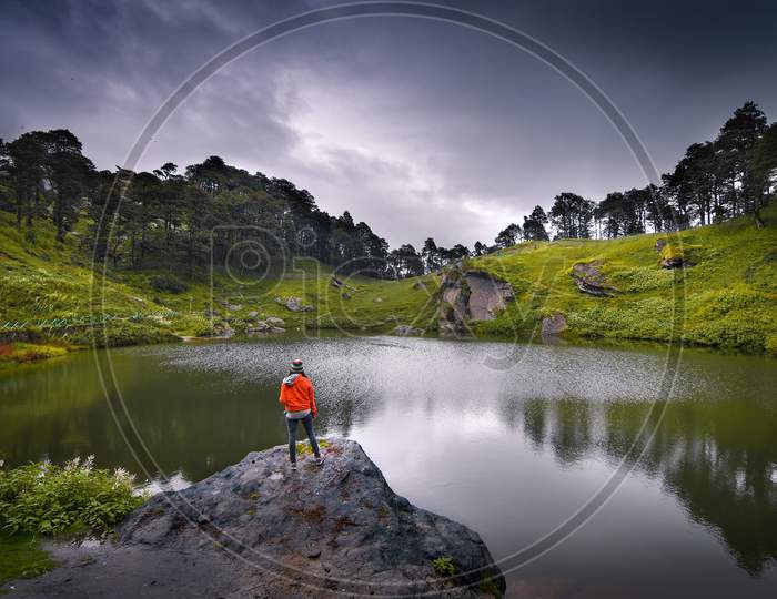 A Landscape Of A Small Lake In Between The Mountain Ranges With A Man Standing Beside It.