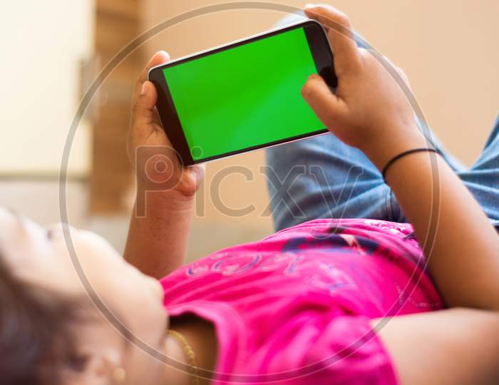 Little Indian Kid With Phone In Her Hands Sleeping On Bed, Mock Up With Green Screen, Focus On Phone.
