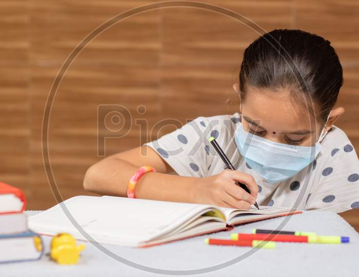 Concept Of Homeschooling, Young Girl With Medical Mask Busy In Writing At Home During Covid-19 Or Coronavirus Pandemic Lock Down.