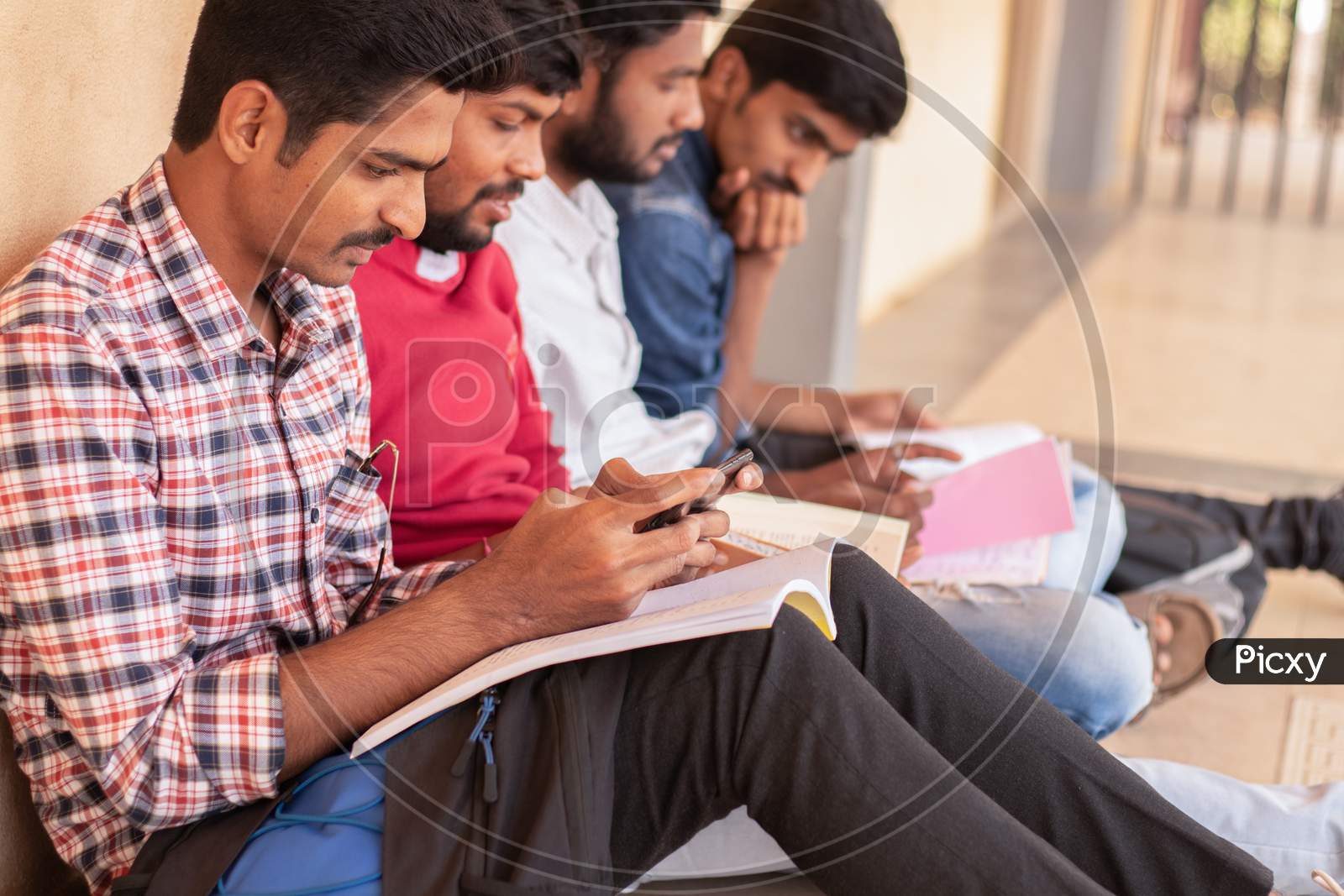 A student using a mobile phone while others are studying by Looking Into Books At College - Education, Learning Student, People Concept