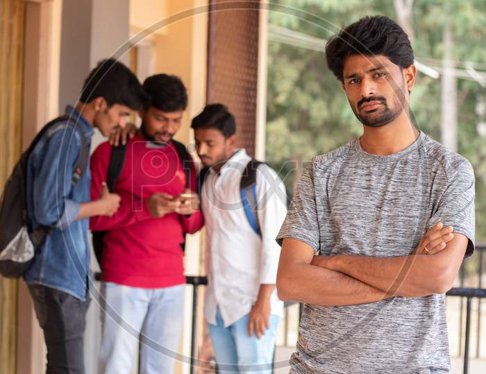 A Young Indian Student Standing and posing towards the Camera At A Campus while others using Mobile in the Background