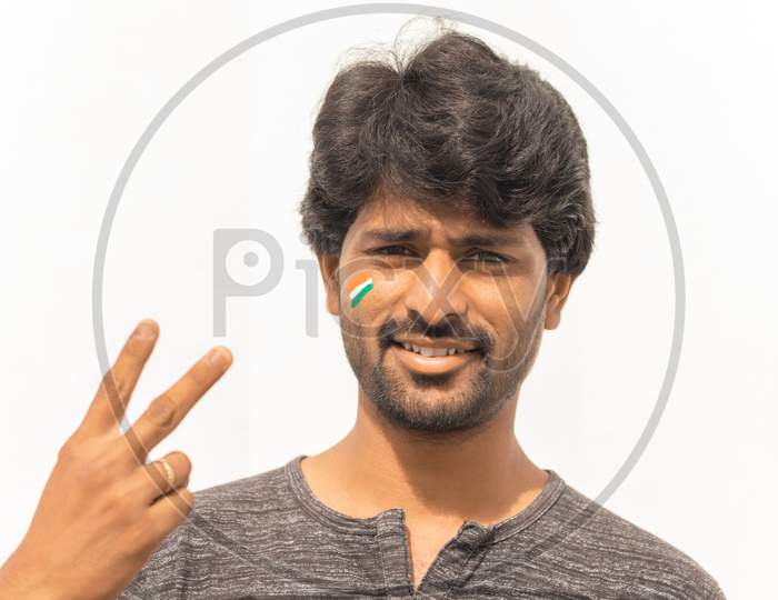 Portrait Of Young Cheerful Indian Male Cricket Sports Fan Painted Indian Flag On His Face And Showing Hand Gesture Of Victory