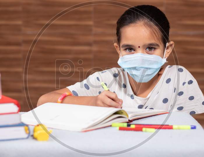 Concept Of Homeschooling And E-Learning, Young Girl With Medical Mask Writing By Looking Into Laptop During Covid-19 Or Coronavirus Pandemic LockDown.