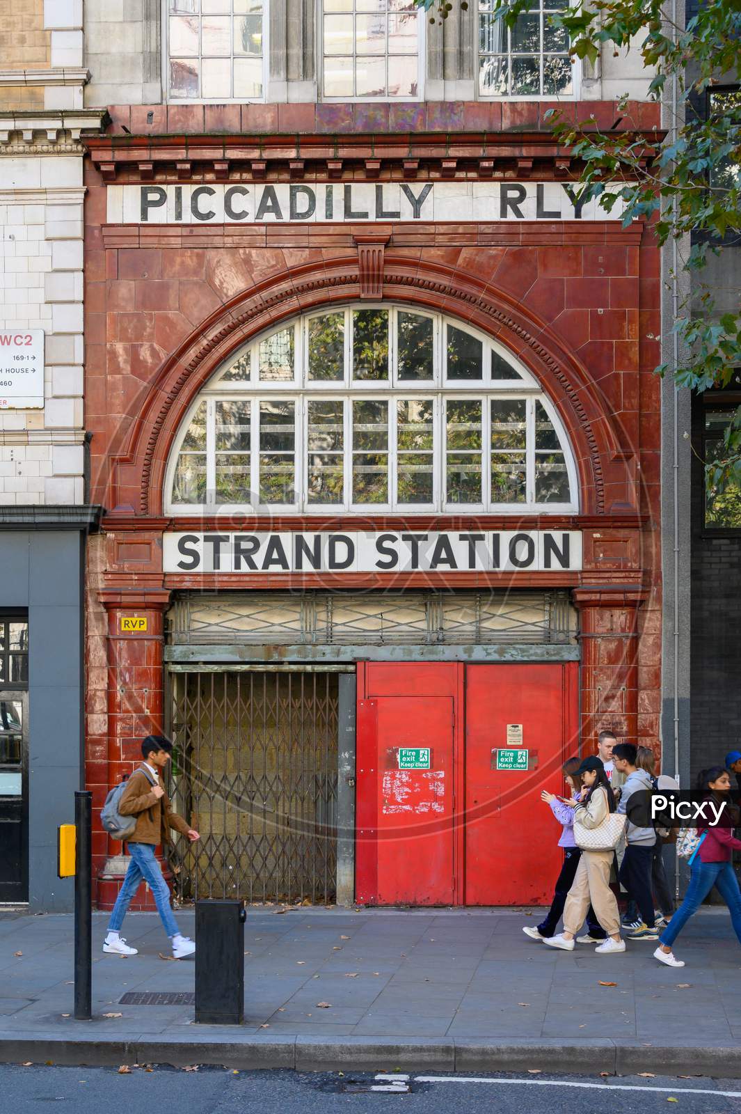 Students Passing In Front Of The Entrance To The Disused London Underground Aldwych Tube Station On The Strand
