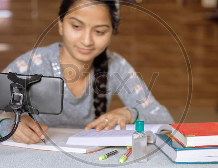 Selective Focus Of Mobile, Concept Of E-Learning At Home Due To Covid-19 Or Coronavirus Isolation - Young College Girl Taking Notes By Looking Into Virtual Class On Mobile Due To Isolation.