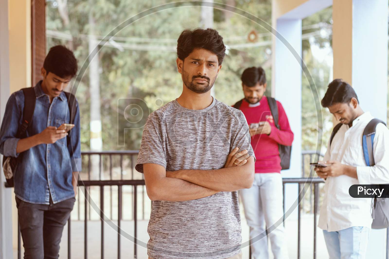 A Young Indian Student Standing and posing towards the Camera At A University Campus while others using Mobile in the Background