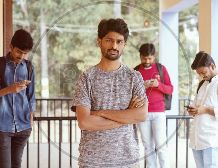 A Young Indian Student Standing and posing towards the Camera At A University Campus while others using Mobile in the Background