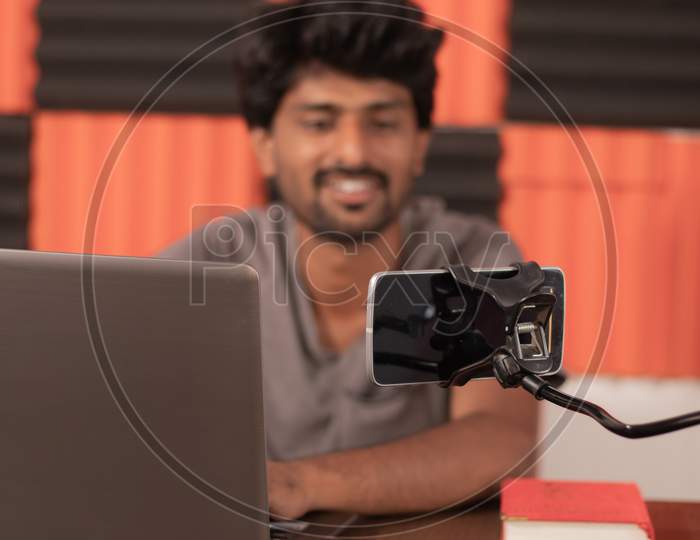 Young Man Looking On Mobile Busy In Video Chat With Friends At Home During Covid-19 Or Coronavirus Pandemic - Smiling Blogger With Laptop Filming Content For Subscribers On Smart Phone Concept