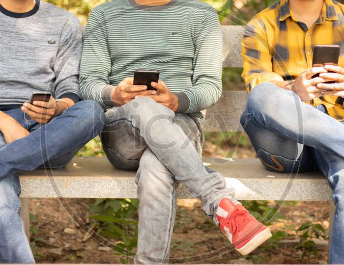 Young Men Busy On Mobile While Sitting On Table At Park - Concept Of Young Generation Addicted To Smartphone, Technology And Electronic Gadgets - Concept Of Modern Youth Lifestyle, Social Interaction.