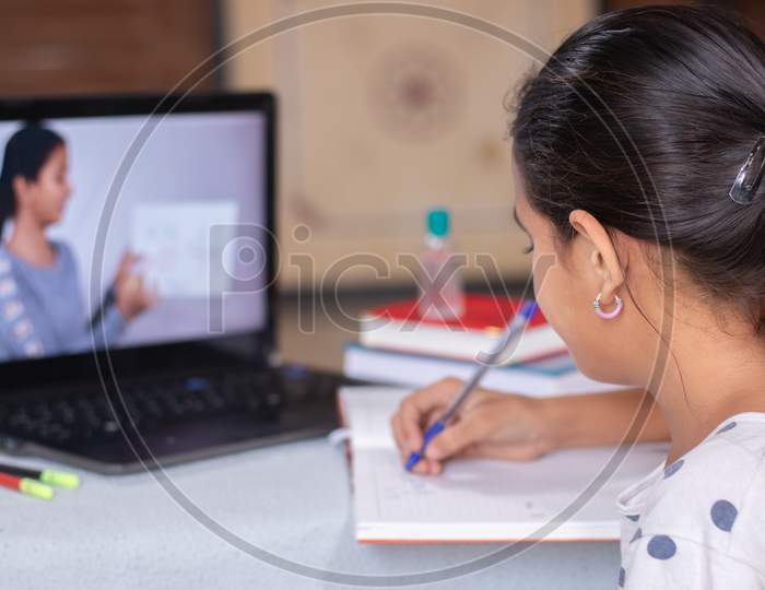 Concept Of Homeschooling Or E-Learning, Young Girl Busy In Writing By Looking Into Laptop While Teacher Explaining During Covid-19 Or Coronavirus Pandemic Crisis.