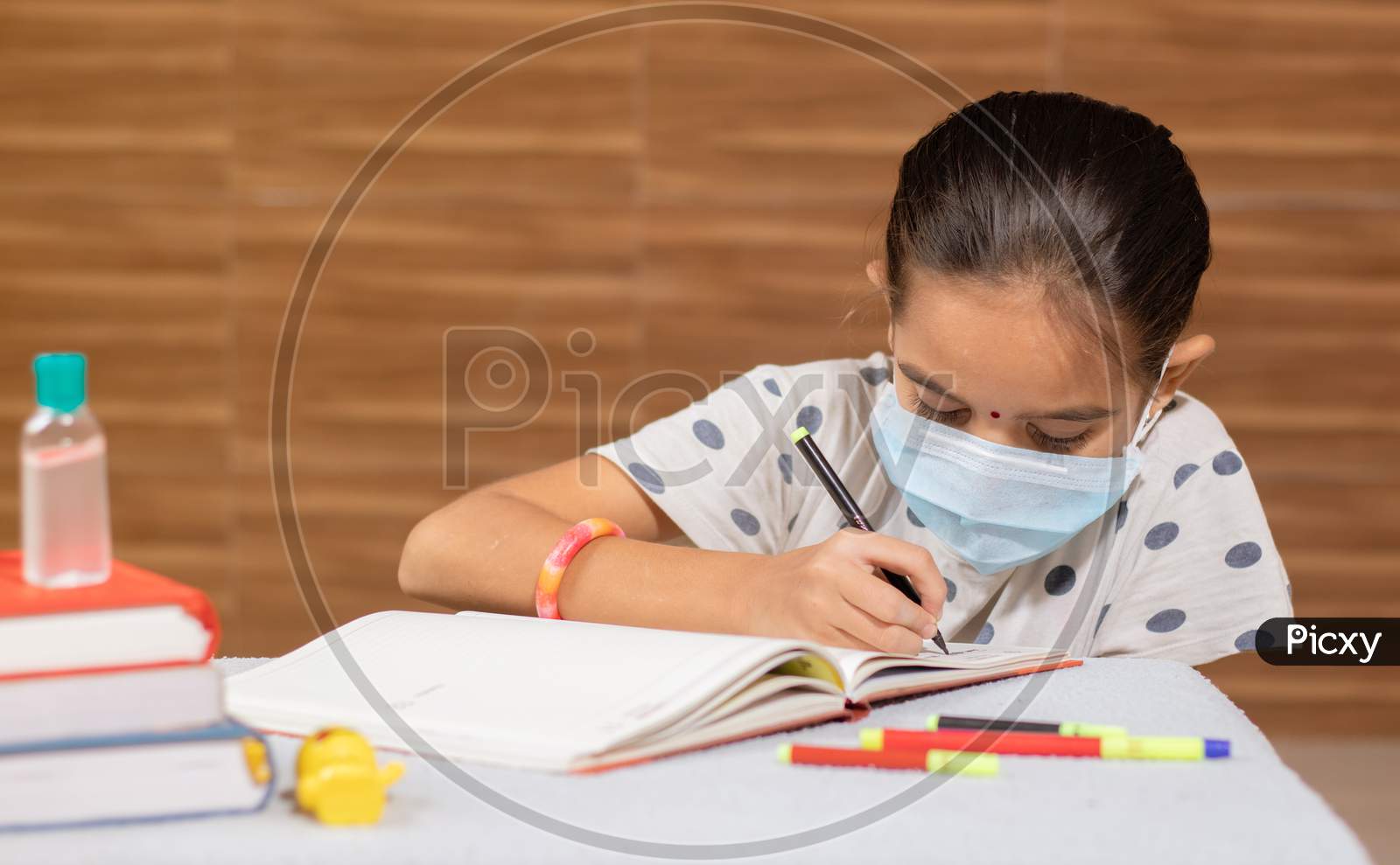 Concept Of Homeschooling, Young Girl With Medical Mask Busy In Writing At Home During Covid-19 Or Coronavirus Pandemic Lock Down.