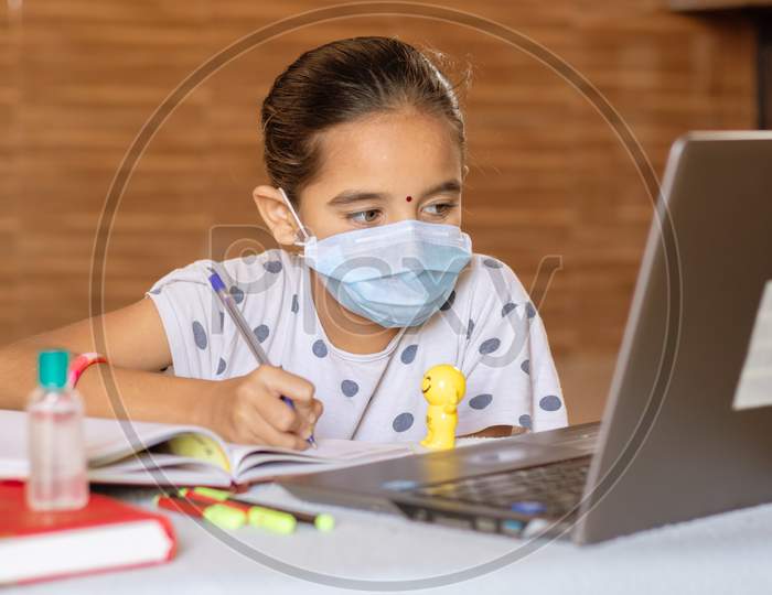 Concept Of Homeschooling And E-Learning, Young Girl With Medical Mask Writing By Looking Into Laptop During Covid-19 Or Coronavirus Pandemic Lock Down.