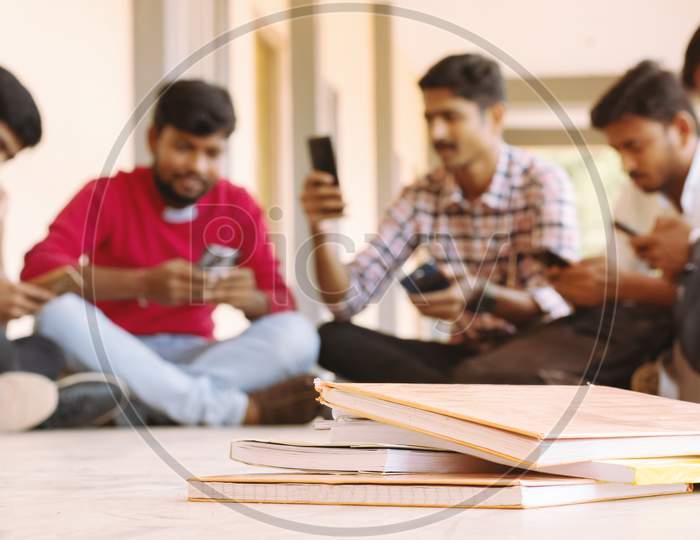 Selective Focus Of Books, Group Of Students Busy On Mobile By Avoiding Books During Exams At College - Teenager Students On Smartphone Video Game Addiction Concept.