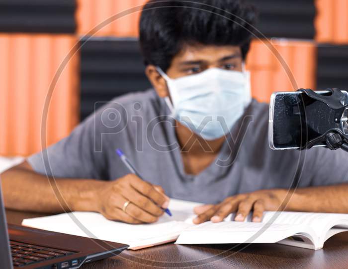Young Man With Medical Mask E-Learning At Home Due To Covid-19 Or Coronavirus Isolation Concept - College Student Taking Notes By Looking Into Virtual Class On Mobile Due To Quarantine.